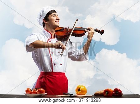 Young Male Chef With Violin Standing Near Cooking Table With Vegetables. Handsome Chef In White Hat 