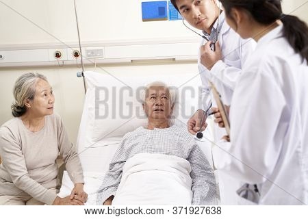 Young Asian Doctor Examining Elderly Patient Discussing His Medical Condition In Hospital Ward