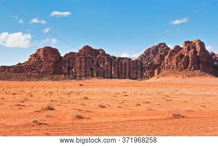 Red Dusty Desert With Large Rock Massif And Blue Sky In Background, Small Off Road Vehicle In Foregr