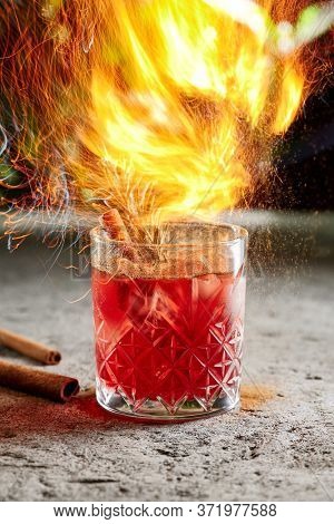 Fiero cocktail with whiskey and cinnamon stick with rosemary twig side view. Alcohol beverage close up with cinnamon powder. Night club drink with ice cubes in bar glassware on blurred background