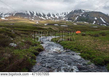 Orange Tent Near Clear Water Stream With View To Mountain Range Among Low Rainy Clouds. Dramatic Alp