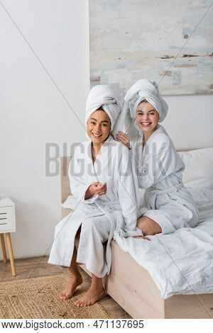 Happy Interracial Women In White Soft Robes And Towels Looking At Camera While Sitting On Bed.