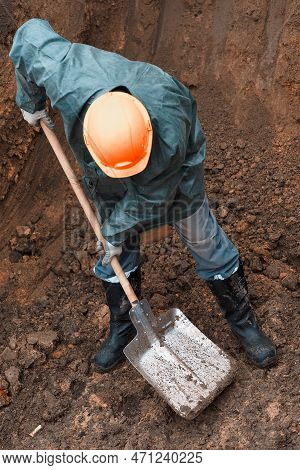 Man In Helmet, Raincoat And Work Clothes With Shovel In Pit Digs Hole. Moil. Rescue Operation. Volun