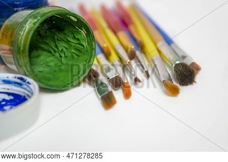 Watercolor And Oil Paints On A White Table. Jars Of Colored Paint Stand Next To The Brushes. For Art