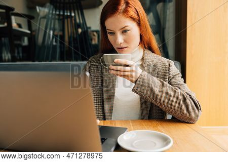 Young Woman With Ginger Hair Holding A Cup Of Coffee And Working On A Laptop In Cafe