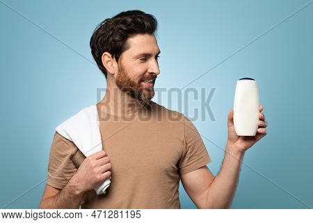 Handsome Bearded Man With Towel On Shoulder Holding And Looking At Shampoo Bottle, Posing Over Blue 