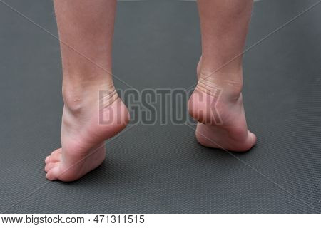 Child Is Barefoot On Mat. Child Does An Exercise For Feet, Standing On Tiptoes. Back View. Preventio
