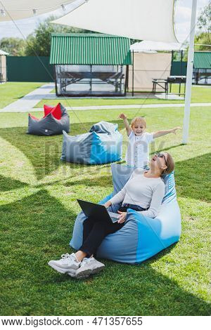 Mother With Laptop, Her Son Playing Outdoors On Green Lawn. Rest And Work With A Child In The Park
