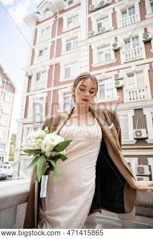 Young Woman In Wedding Dress And Beige Blazer Holding Bouquet Of Flowers.