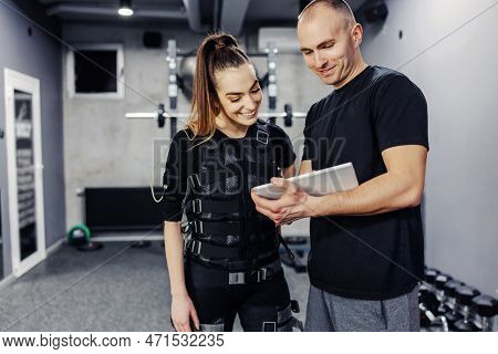 A Woman In A Ems Suit And A Male Trainer In Sportswear Holding A Tablet. They Stand In The Middle Of