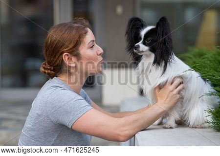 Caucasian Red-haired Woman Kissing Pappilion Dog Outdoors. Black And White Continental Spaniel.