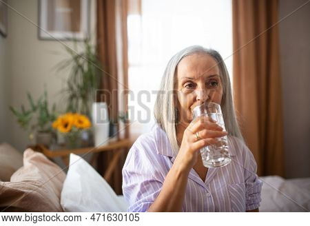 Portrait Of Senior Woman Sitting On The Bed In Pajamas And Drinking Water.