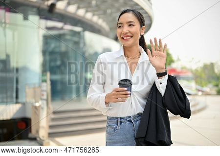 Cheerful Asian Businesswoman Walking On The Street, Waving Hand, Saying Hi To Her Colleagues