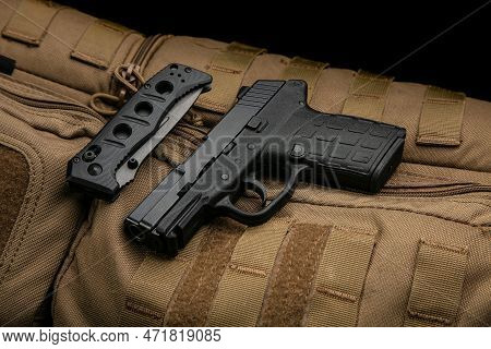 A Small Modern Pistol And Penknife On A Brown Military Backpack. Self Defense Kit. Dark Background.