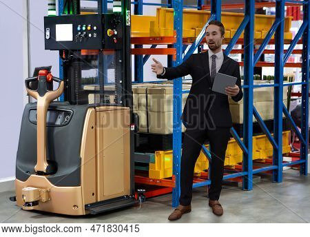 Businessman Stands In Front Of Agv Forklift In Modern Warehouse. Manager Use Tablet To Check Shippin