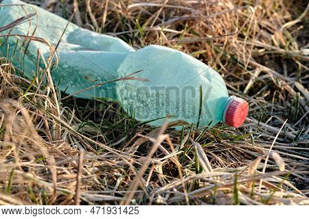 Crumpled plastic bottle covered with ice and snow in frozen dry grass. Green with red bottle cap. Pl