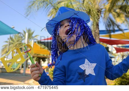 Carnival, Purim In Israel. A Cheerful Little Boy In A Hat, Funny Glasses, A Wig With A Rattle, Raash
