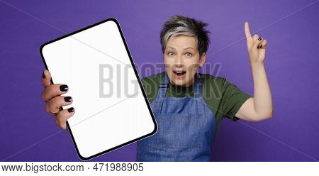 Surprised Mature 50s Woman Wearing Denime Apron Holding Bif Mock Up Tablet Isolated On Violet Studio