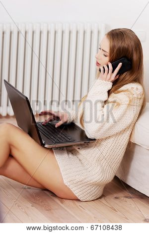 Beautiful casual woman in brught sweater working on laptop and talking through phone. She's sitting on the wooden floor.