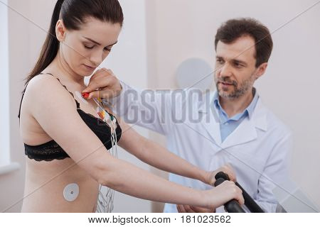 Healthy cardio. Distinguished smart contemporary cardiologist connecting vires to several sensors on womans chest while testing modern equipment for indicating cardiac diseases