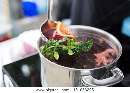 Making mulled wine for sale at country fair. Glintwine with spices, greens, citrus and cloves in big metal stock pot, mixed with soup ladle.