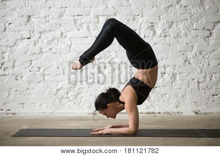 Mid aged yogi attractive woman practicing yoga concept, standing in vrischikasana exercise, Scorpion pose, working out, wearing sportswear, black tank top and pants, full length, white loft background