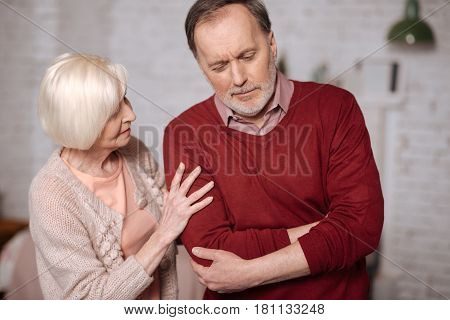 Is it appendix. Portrait of elderly man standing and touching his aching stomach area while his loving wife giving support to him.