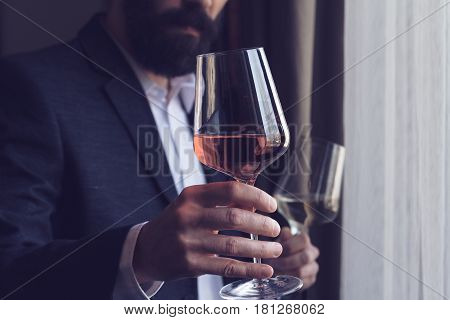horizontal close up of a Caucasian man with beard black suit and white shirt offering a tall glass of rose wine at an event by the window natural light