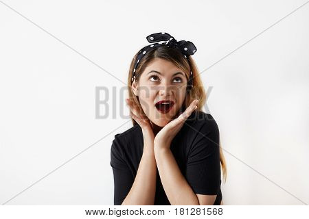 Portrait of beautiful young surprised woman dressed in pin-up style holds cheeks by hand and looks up. Caucasian blond model screaming Wow posing in vintage concept. Expressive facial expressions