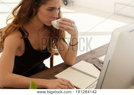 Working process photo. Cropped image of attractive female writer drinking coffee while searching information for new article about ecological situation via contemporary computer in the workplace