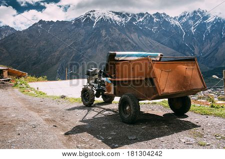 Stepantsminda Gergeti, Georgia. Small Old Two-wheel Tractor Or Walking Tractor French With Trailer Used In A Rural Household. Tractor On Road In Village On Mountain Background