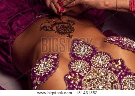White woman a mehendi artist paints an ornament of henna on an eastern beautiful girl's stomach