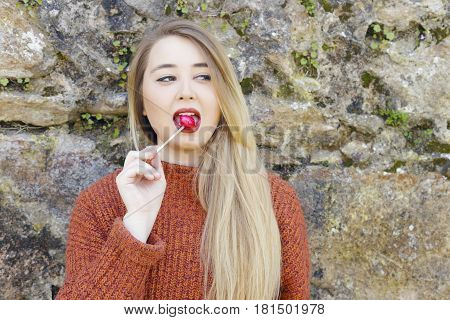 Cheerful young girl on orange sweater with candy on stick Chupa Chups outdoors on a sunny day against a stone wall.