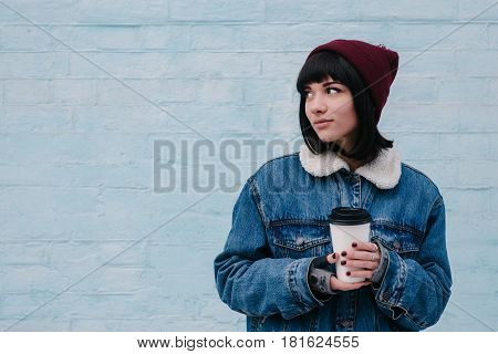Young hipster girl in a blue denim jacket and hot drinks in hand on light background blaktytnomu looks aside and smiles