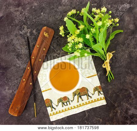 Green herbal tea in a cup incense stick and holder napkin with elephants bright bouquet of linden tree flowers. Oriental composition on dark stone background. Flat lay top view
