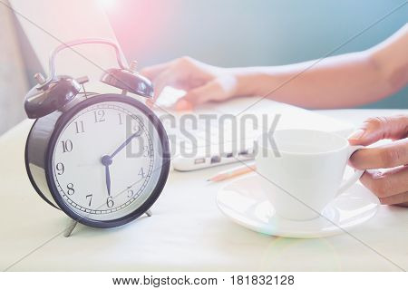 Woman working on laptop computer and drinking coffee on modern white working space with classic alarmclock