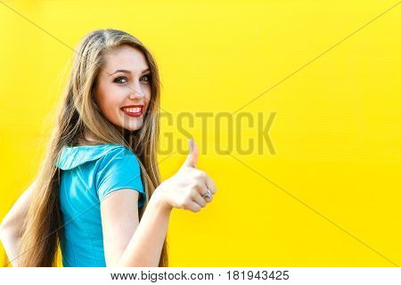beautiful blonde girl in blue dress showing finger up sign OK on a yellow background.
