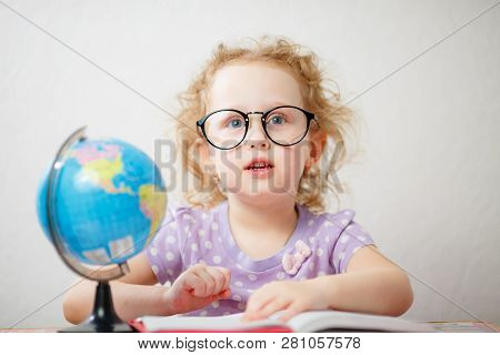 Close Up Photo Portrait Of Funny Little Girl With Glasses. Shes Leafing Through The Book On The Tabl