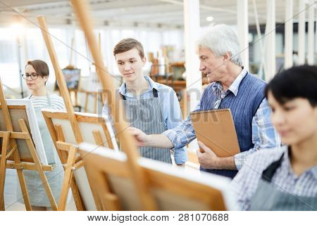 Portrait Of Mature Art Teacher Helping Group Of Students Painting Picture On Easels In Art Class, Co