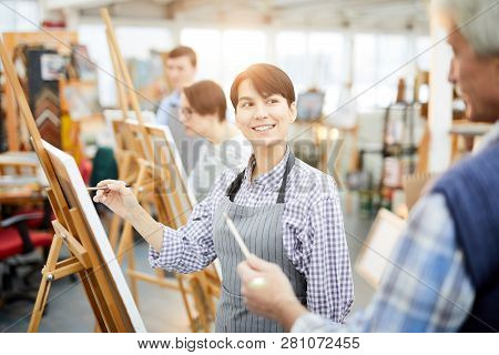 Waist Up Portrait Of Smiling Young Woman Painting Picture On Easel In Art Class And Looking At Teach