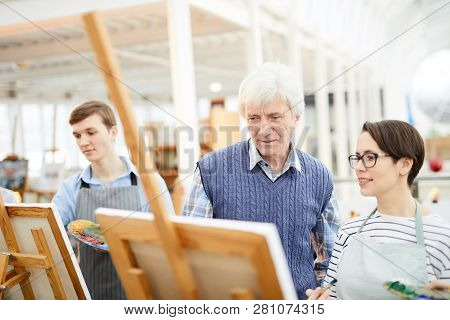 Portrait Of Mature Art Teacher Helping Group Of Students Painting Pictures On Easels In Art Class, C