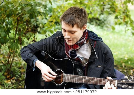 Young Guy Sitting In The Park And Playing The Black Guitar
