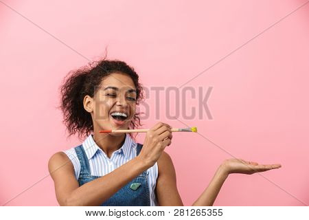 Beautiful young african woman atrist holding a paint brush over pink background, presenting copy space