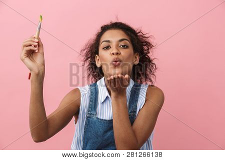 Beautiful young african woman atrist holding a paint brush over pink background, sending kiss