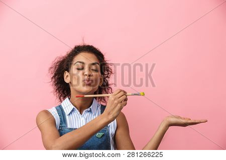 Beautiful young african woman atrist holding a paint brush over pink background, presenting copy space