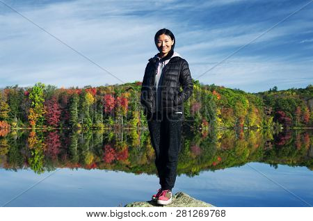 A Chinese Woman Standing On A Rock Looking At The Fall Foliage Reflecting In The Water While Sitting