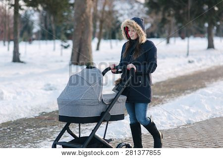Beautiful Mother Walking In The Park With Her Little Baby In Stoller. Woman Dressed In Blue Jaket Wi