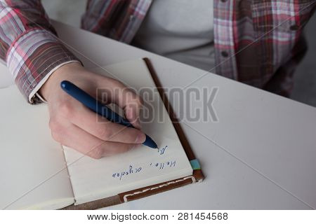 Close Up Shot Of Male Hand Writing In Notebook. Hand Of Young Male Wearing Long Sleeved Button Up Sh