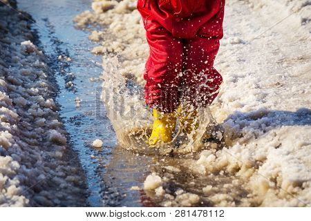 Kid Legs In Yellow Rainboots Jumping In The Ice Puddle With Melting Snow At Sunny Spring Day, Outdoo