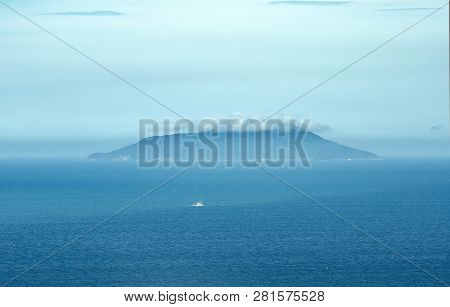 Small Island In The Middle Of The Sea On A Cloudy Day, Clouds Very Low And Fog. Some Boats Navigatin